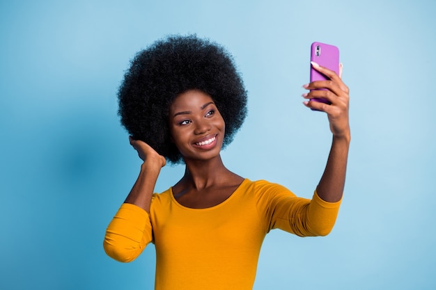 Fotoportret van mooi zwart gevild meisje dat selfie glimlachend ontroerend kapsel neemt geïsoleerd op een levendige blauwe kleur achtergrond