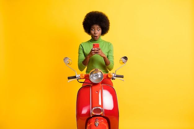 Fotoportret van opgewonden motorrijder op motorfiets met mobiele telefoon in twee handen met een casual groene trui geïsoleerd op een levendige geel gekleurde achtergrond