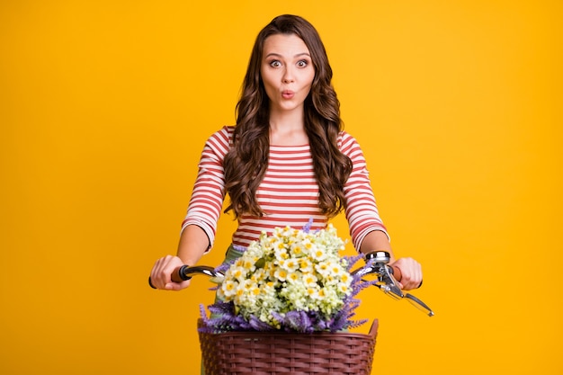 Fotoportret van verrast meisje dat handen op de fiets houdt met een mand met wilde bloemen geïsoleerd op een felgele achtergrond