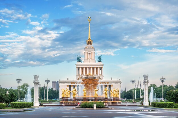Фонтан Дружбы народов с золотыми статуями девушек и Центральный павильон с высоким шпилем на территории ВДНХ в Москве летним вечером