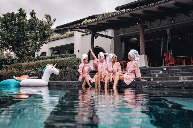 Четыре очаровательных скинну и милые девушки-модели в одинаковой одежде позируют и сидят у бассейна с надувными игрушками на фоне виллы и домов. Концепция расслабления