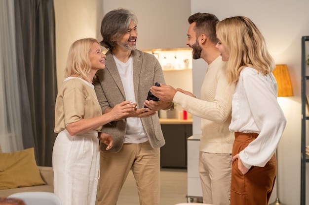 Four cheerful family members discussing sort of wine held by young man while standing in front of camera and laughing during conversartion