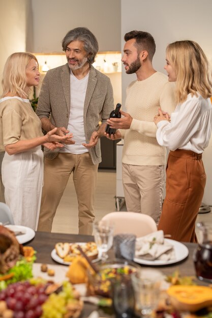 Four cheerful family members discussing wine held by young man while standing in front of served festive table with tasty homemade food