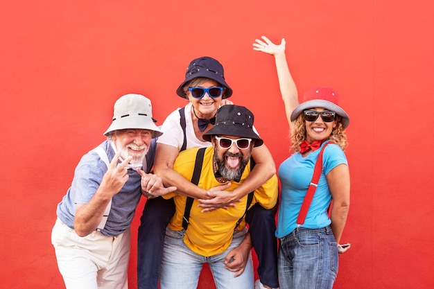 Four cheerful people standing in front of a red wall. Gesturing while the son carries on his shoulders the mom. Large smiles and colorful dresses with suspenders and bow ties. Complicity and happiness