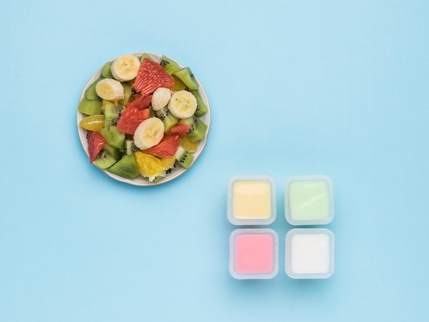 Four cups of yogurt and a bowl of fruit salad on a blue background. Milk and fruit food. Flat lay.