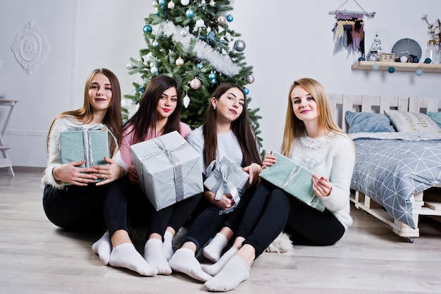 Four cute friends girls wear on warm sweaters black pants against new year tree with christmas decoration at white room holding gift boxes at hands