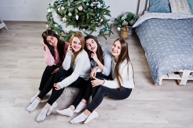 Four cute friends girls wear on warm sweaters, black pants against new year tree with christmas decoration at white room.