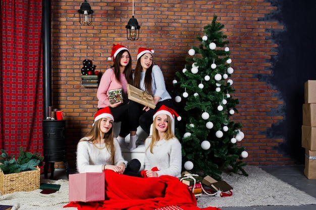 Four cute friends girls wear on warm sweaters black pants and santa hats against new year tree with christmas decoration at studio