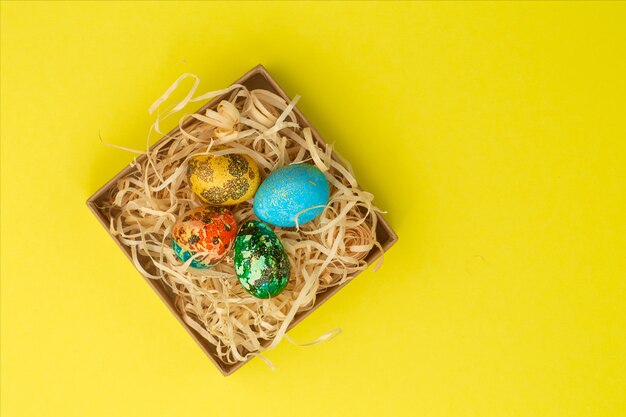 Four Easter eggs painted in red, blue, green and yellow, lies in a box, inside which are wooden shavings. Painted Easter eggs on a yellow background. Flat lay . Copy space