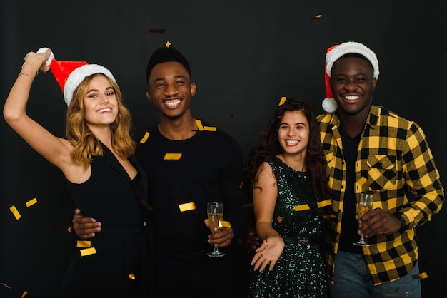 Four friends of different nationalities throw confetti in santa claus hats and antlers and drink champagne against a black background.