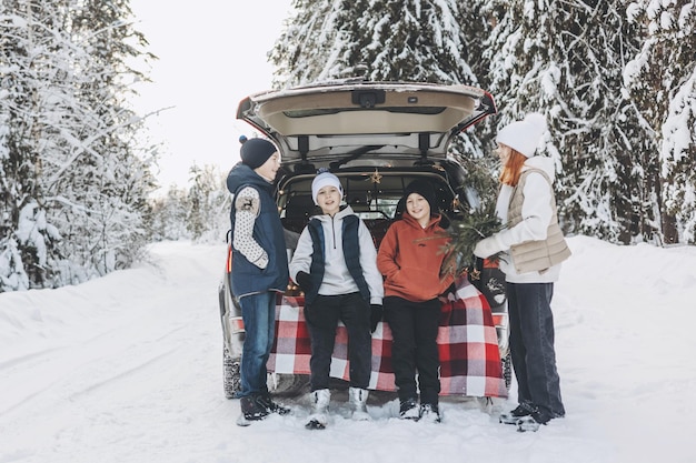 Four friends teenagers boys and girl with thermos sitting in trunk of car decorated for Christmas and New year Winter picnic in snowy forest Road trip and local travel Friends hanging out together