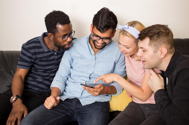 Four friends wearing casual clothes sitting on couch at home, using mobile phone together, posting on social networks and surfing the net on smartphone.