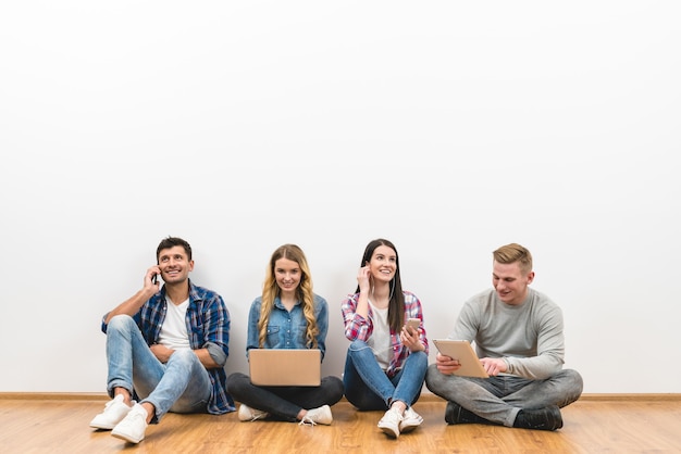 The four friends with gadgets sit on the floor on the white wall background