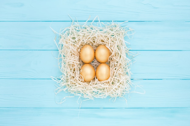 Four golden Easter eggs in square nest on blue background, flat lay.