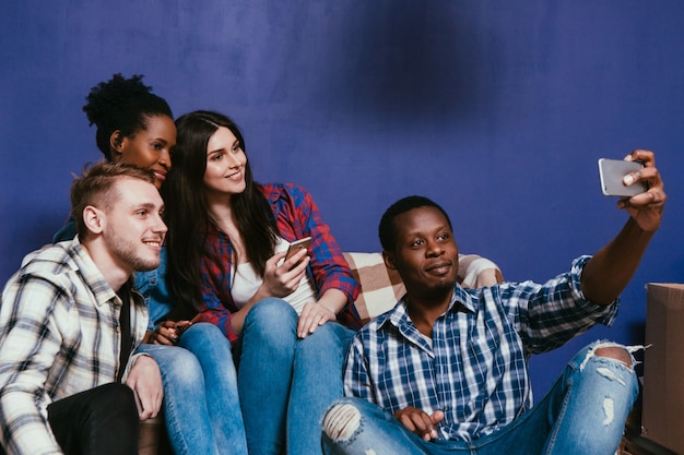 Four happy young friends make group selfie with mobile on home couch