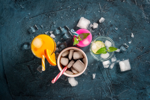 Four kinds of refreshing drinks with ice on a dark blue background and ice cubes. Concept night club, night life, party, thirst. Orange, mint and cucumber, strawberry, cola. Flat lay, top view