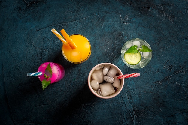 Four kinds of refreshing drinks with ice on a dark blue background and ice cubes. Concept night club, night life, party, thirst. Orange, mint and cucumber, strawberry, cola. Flat lay, top view