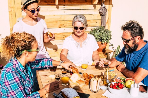 Quattro persone si godono la colazione in terrazza sotto la luce del sole. genitori con figlio adolescente e nonna. tavolo in legno con torte fatte in casa, frutta e caffè