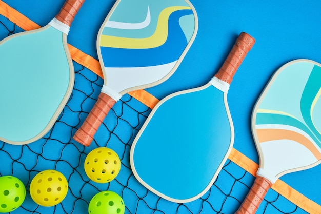 Photo four pickleball paddles in blue tones on top of a net on a blue background with three balls next to it