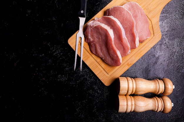 Four pieces of raw steaks meat on wooden board next to two salt and pepper shakers on black wooden backgorund