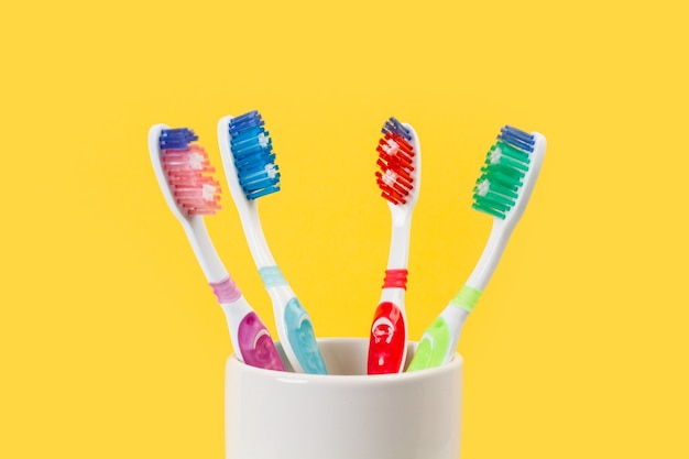 Four plastic colored toothbrushes in a ceramic glass on a yellow background