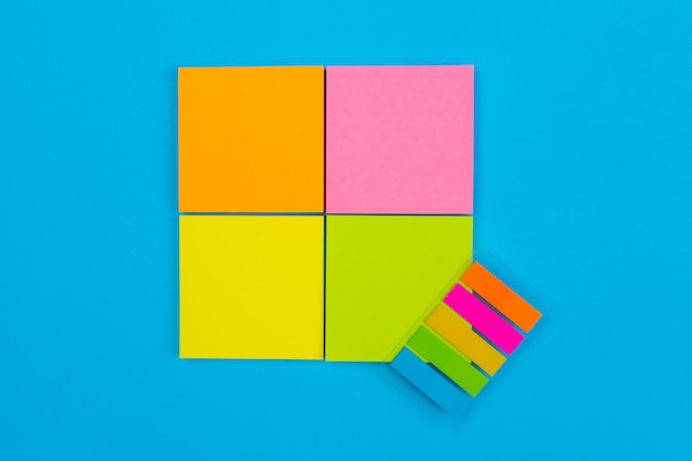 Four small sets of yellow, orange, pink, green stickers folded in the form of a square on a blue  on one of which are stickers in the form of stripes. Close-up. Place for notes.