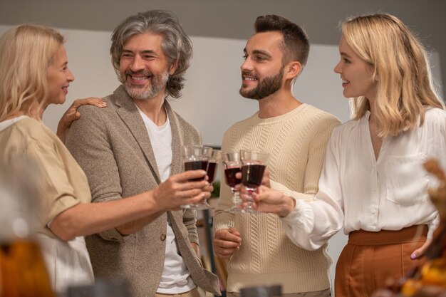Четыре улыбающихся члена семьи в повседневной одежде, чокаясь с бокалами красного вина и смотрящие на счастливую зрелую белокурую женщину во время празднования