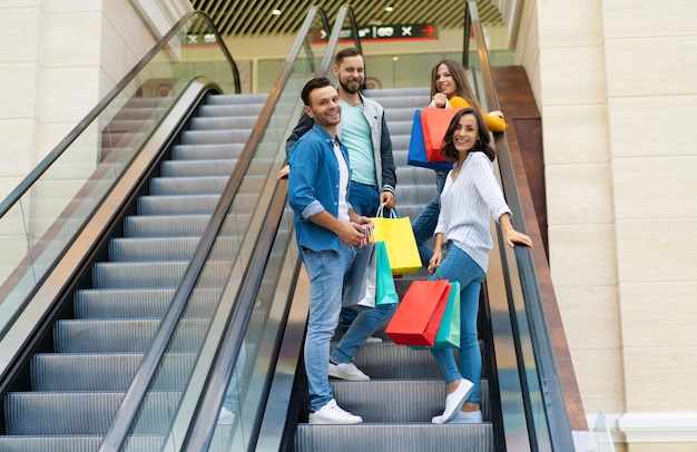Four stylish smiling excited beautiful modern friends in casual clothes with paper bags are having fun while standing on the escalator in the mall during shopping.