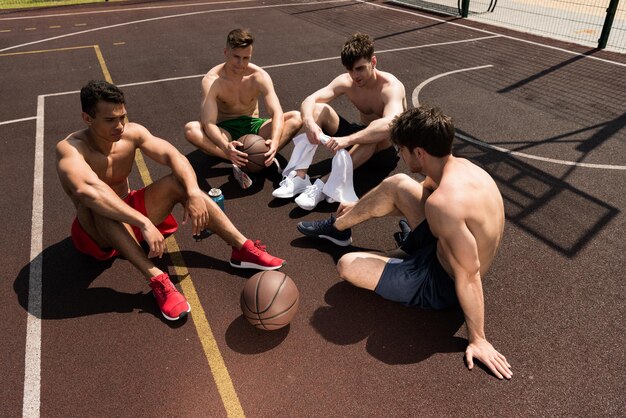 Photo four tired shirtless basketball players sitting at basketball court in sunny day