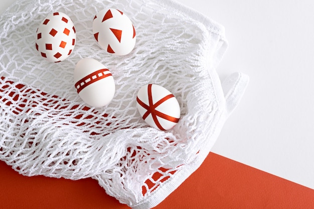 Four white Easter eggs with red geometric pattern on white mesh eco bag on red and white background, easter concept