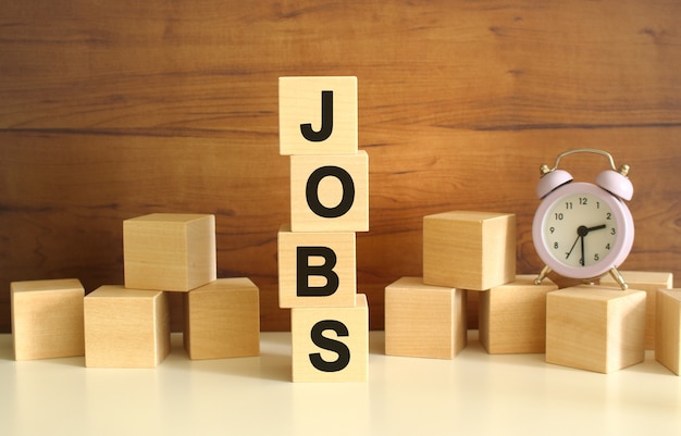 Four wooden cubes stacked vertically on a brown background form the word JOBS. Cubes are scattered nearby and there is a clock. Front View Concepts