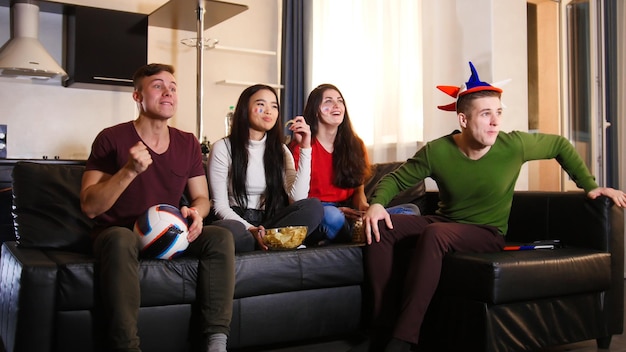 Four young people sitting on the sofa and watching football match wearing russian attributes