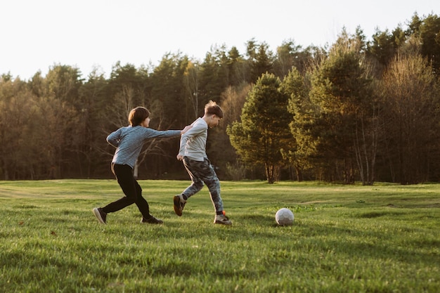 foxy teenager in gray grabbing friend on clothes attempting to overtake boy throughout running towards white ball competitive teens playing football on green field near forest and enjoying game