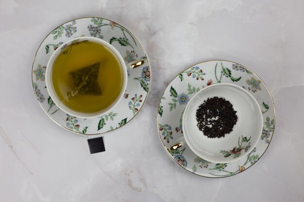 Foto tazze e piattini frazionari con una bustina di tè verde e foglie di tè nero su un tavolo bianco