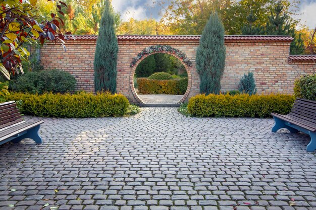 A fragment of landscape design in an autumn park with beautifully trimmed trees and bushes an arch and a paved alley