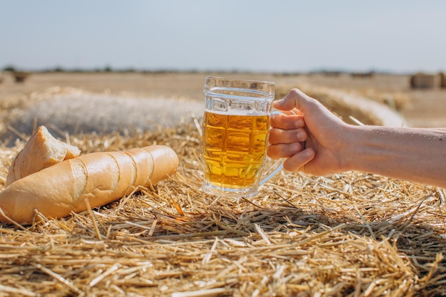 パンと小麦畑の背景に新鮮な冷たいビールのグラスを持っている男性の手の断片