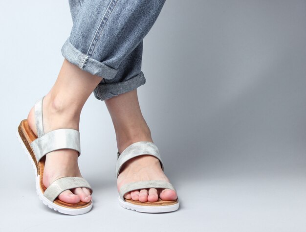 Fragment van vrouwelijke benen in spijkerbroek en trendy lederen sandalen op wit. Stijlvolle zomerschoenen voor dames.