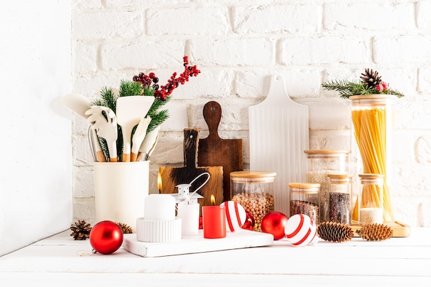 A fragment of a white wooden countertop with various kitchen utensils and Christmas decorations candles balls branches of spruce