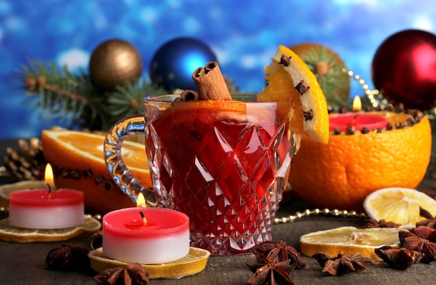 Photo fragrant mulled wine in glass with spices and oranges around on wooden table on blue background