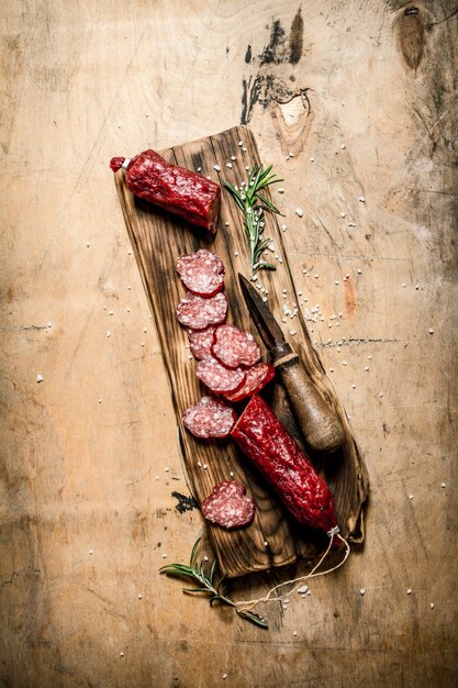 Fragrant salami with rosemary and old knife on wooden table.