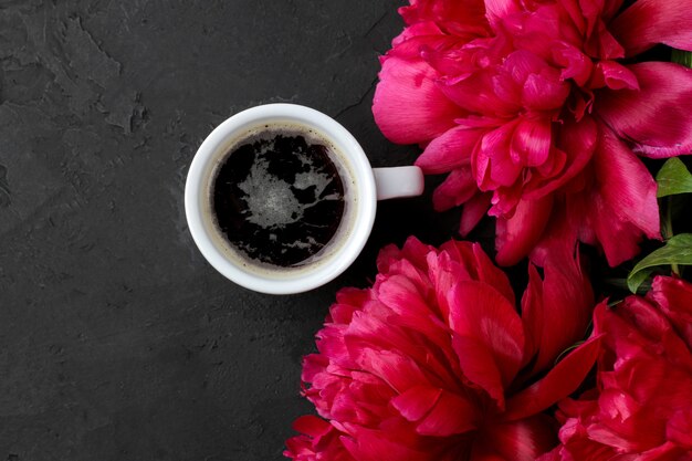 Frame of beautiful bright pink flowers peonies and a cup of coffee on a black graphite background. top view.