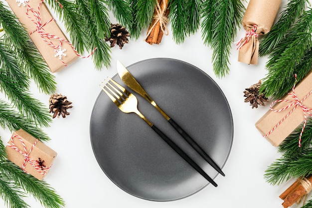 Frame of fir branches cones and gifts for empty black plate and black cutlery on festive table