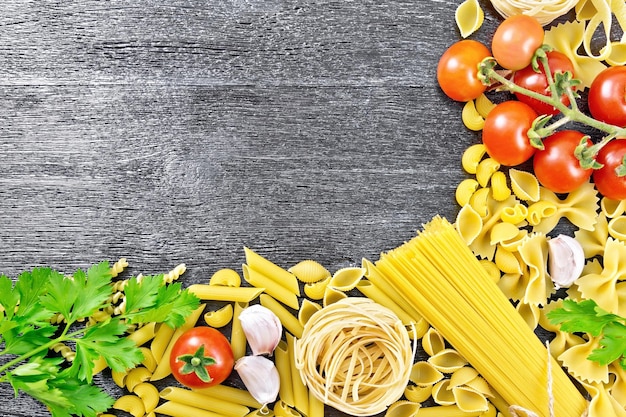 Frame gemaakt van verschillende pasta, tomaten, knoflook en peterselie op een zwarte houten plank achtergrond