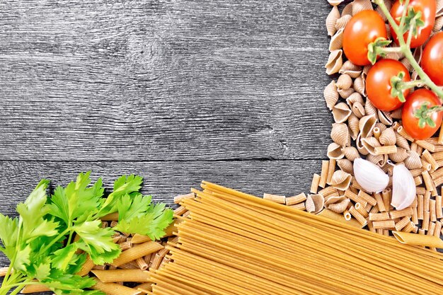 Frame made of assorted rye and wheat whole grain pasta, tomatoes, garlic and parsley on a black wooden board background