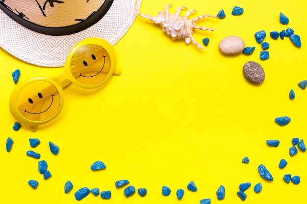 Frame made of sunglasses with a smile, hat, decorative stones and shell on a yellow background. Top view