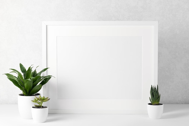 Photo frame mockup with white wooden frame on a white table, houseplants. show text or product.