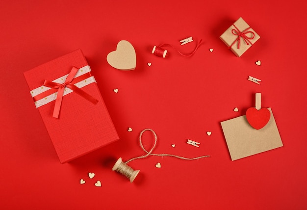 Frame of Valentine gifts, red giftbox, hearts, twine, clothespins and brown paper card on red background, close up flat lay, elevated top view, directly above