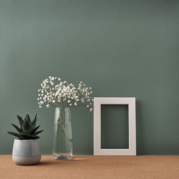 A frame, a vase of white flowers and a house plant in a pot stand on a cork floor