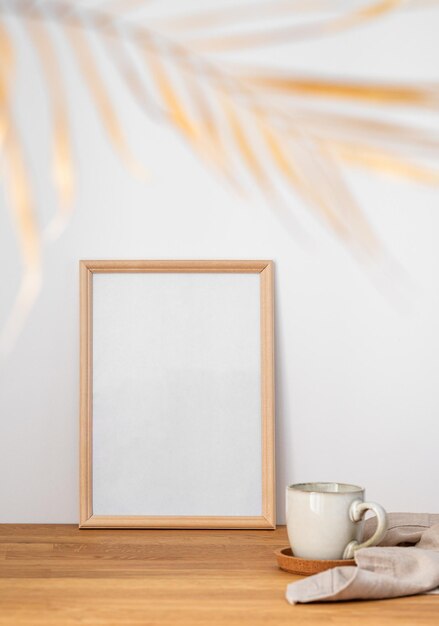 Foto una cornice con una tela vuota contro una parete leggera e su un tavolo di legno con foglia di palma sfocata e una tazza di tè