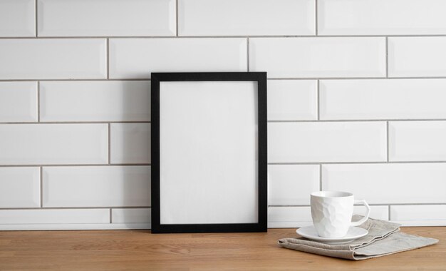 A frame with a blank canvas against a white tile wall and on a wooden tabletop with cup of tea